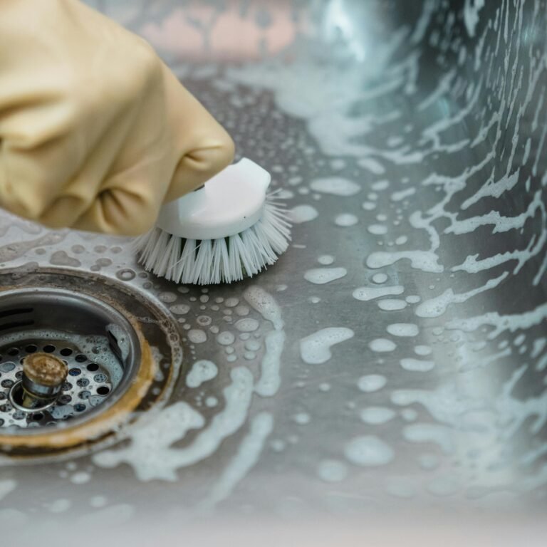 Close-up of a hand in gloves scrubbing a soapy kitchen sink with a brush.